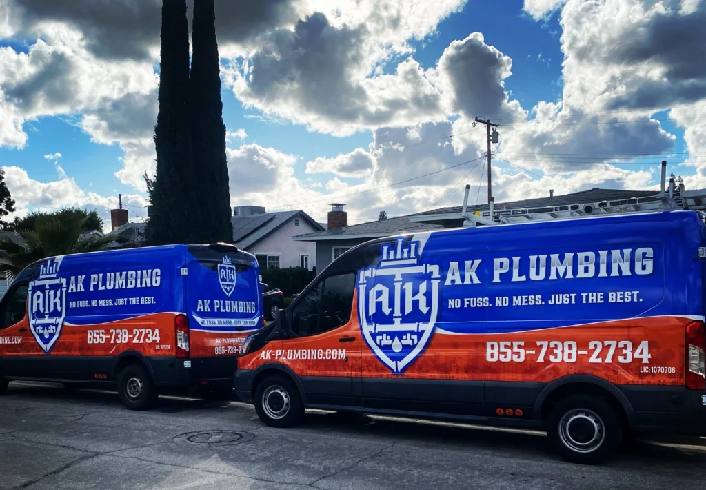 Two AK Plumbing vans parked on a residential street. The vans are blue and orange with company logos and phone number displayed prominently. Cloudy sky and trees in the background.