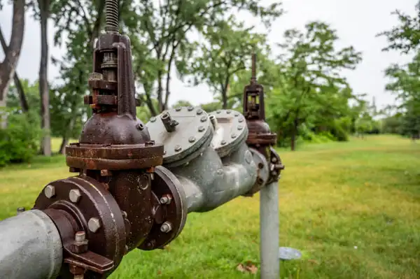 A large pipe with rusted valves in a grassy area, surrounded by trees.
