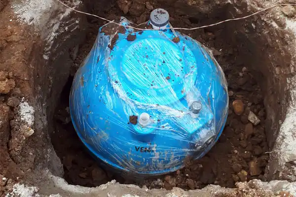 A blue water tank partially buried in a dirt hole, wrapped in protective plastic, surrounded by soil and stones.