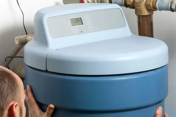 Person adjusting a blue water softener unit with a digital display and brass pipes in the background.