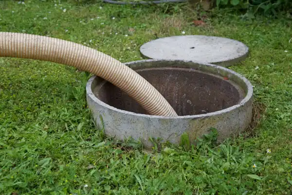 A hose is inserted into an open concrete manhole or septic tank on a grassy area. The round lid is partially visible on the ground nearby.
