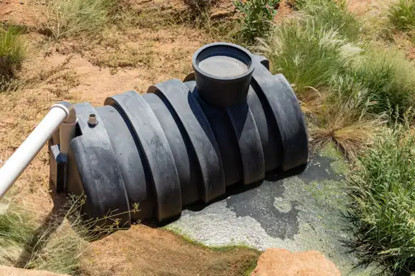 Black plastic septic tank partially buried in the ground with some vegetation around, connected by a white pipe.